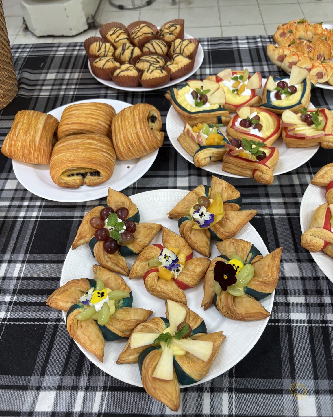 Cận cảnh các đĩa bánh sừng bò, bánh ngọt và bánh pastry trang trí nho, táo và hoa ăn được trên khăn trải bàn kẻ caro.
