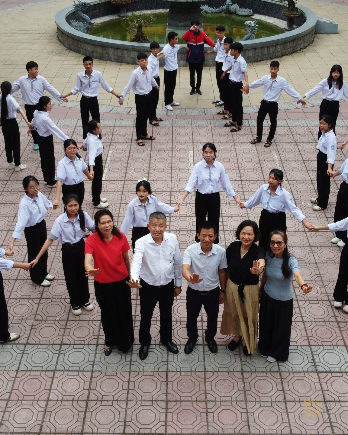 Nhìn từ trên cao: học sinh áo trắng và thầy cô đứng nắm tay nhau thành vòng tròn lớn quanh sân trường lát gạch.