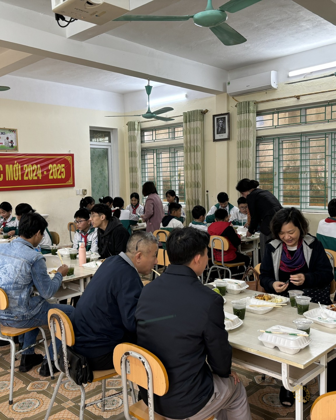 Học sinh THCS Đại An ngồi ăn trưa cùng thầy cô và thành viên Quỹ Hoa Mặt Trời trong phòng học có băng-rôn chào mừng năm học mới.
