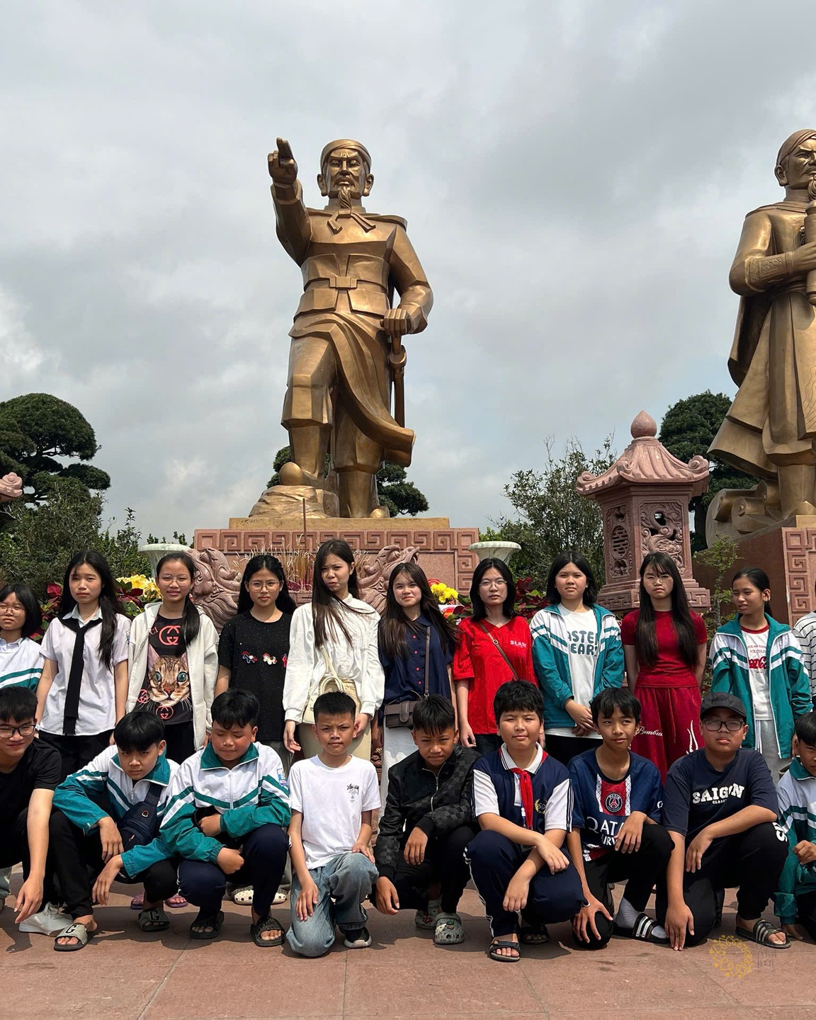 Nhóm học sinh Tân Khánh đứng thành hai hàng trước cụm tượng đồng tại Bạch Đằng Giang, phía sau có băng-rôn vàng ghi "Lễ".