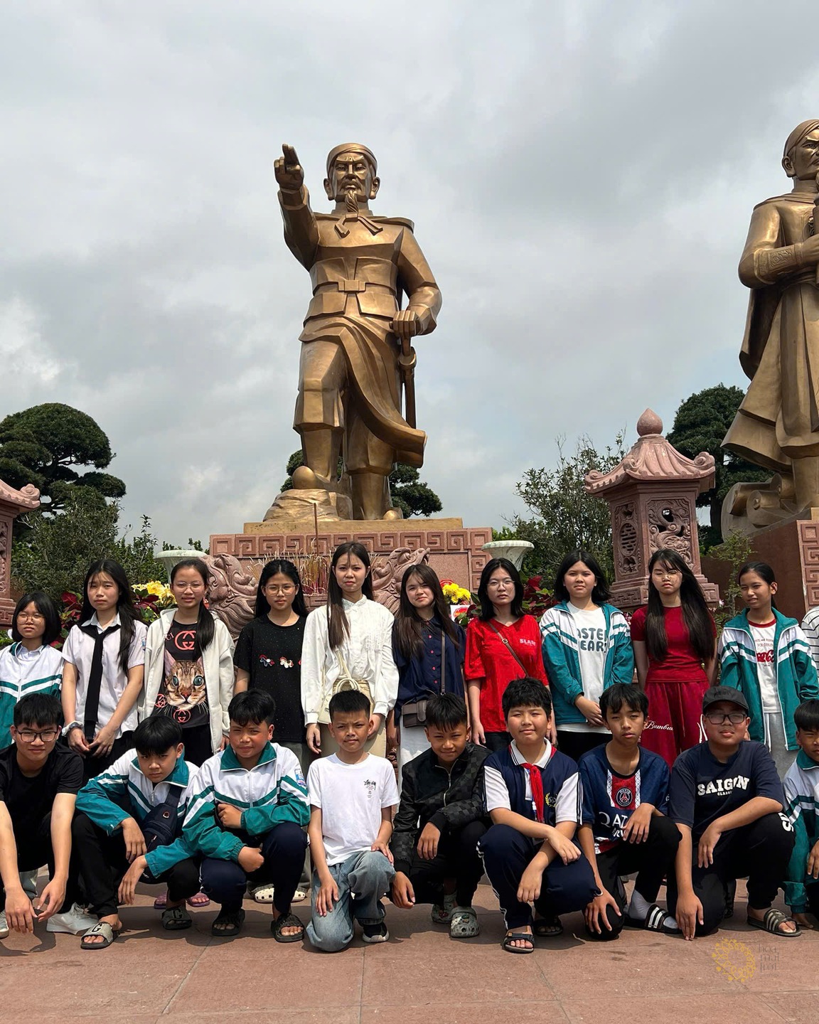 Các em học sinh Trường Tân Khánh tạo dáng chụp ảnh kỷ niệm trước tượng đài ba vị anh hùng dân tộc, trời nhiều mây.