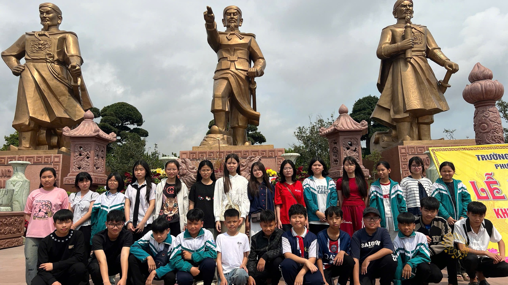 Các em học sinh Trường Tân Khánh tạo dáng chụp ảnh kỷ niệm trước tượng đài ba vị anh hùng dân tộc, trời nhiều mây.