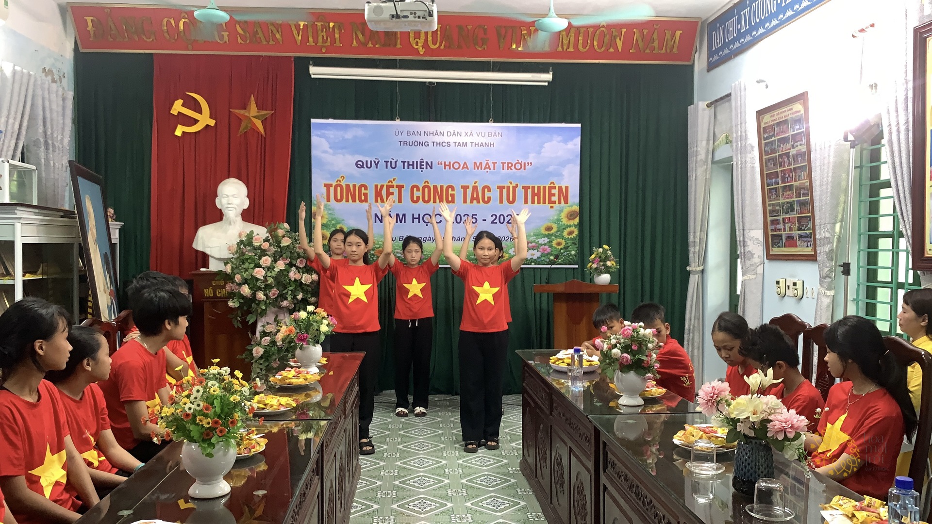 Ba em học sinh mặc áo cờ đỏ sao vàng đứng cạnh nhau trước phông "Tổng kết công tác từ thiện" của Quỹ Hoa Mặt Trời, cùng đưa hai tay lên trong một tiết mục văn nghệ; các bạn cùng lớp ngồi hai bên dãy bàn gỗ theo dõi.