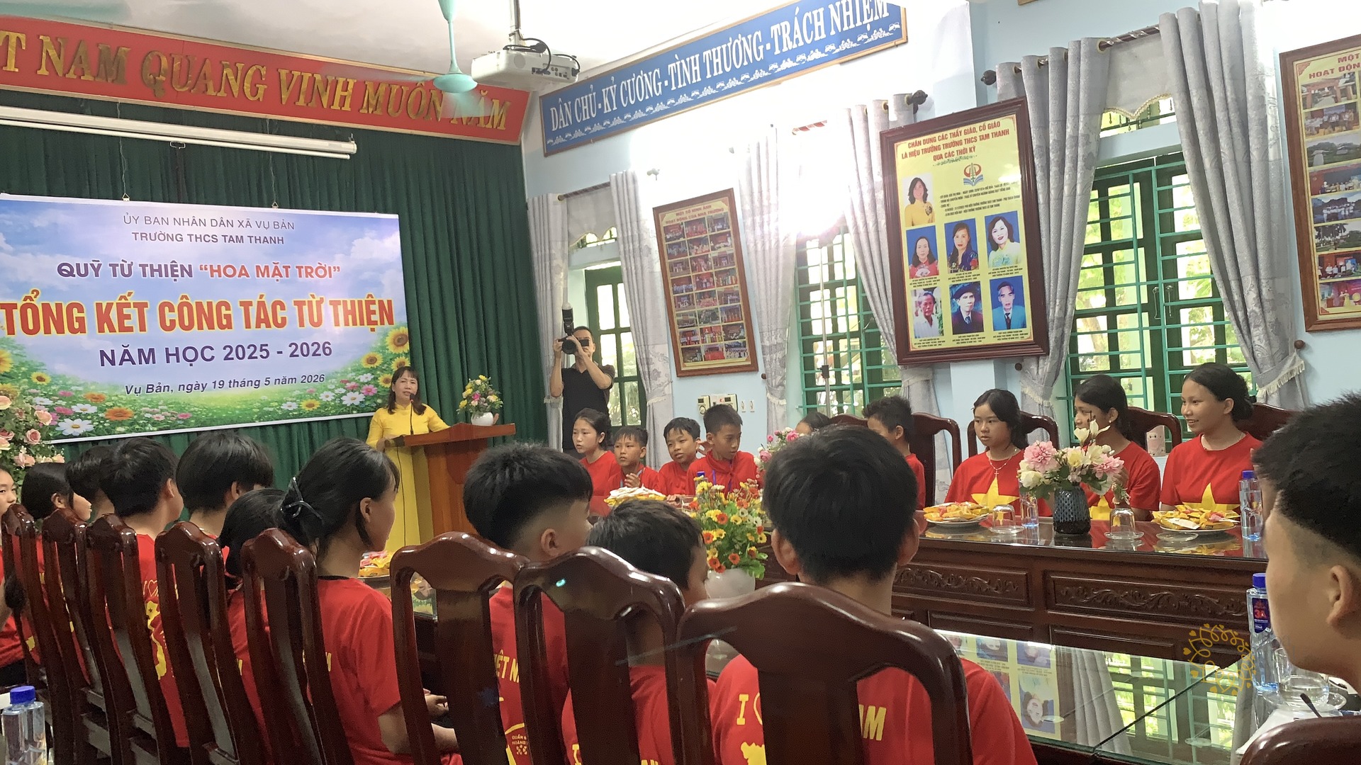 Một cô giáo mặc áo dài vàng đang phát biểu tại bục trong hội trường Trường THCS Tam Thanh; các em học sinh áo đỏ ngồi quây quanh các dãy bàn gỗ phía trước và bên phải.